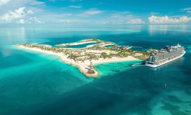 A cruise ship docked beside a small sandy island with a striped lighthouse