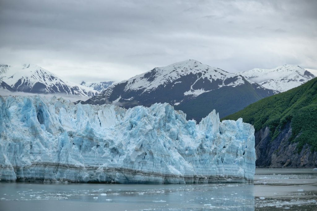 Jagged white-and-blue face of Hubbard Glacier rising from icy water beneath snowy peaks in Alaska